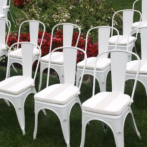 May include: Rows of white metal chairs with curved backs and striped seat cushions are arranged on a green lawn. The chairs are set up for an outdoor event, with red and white flowers in the background.