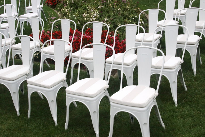 May include: Rows of white metal chairs with curved backs and striped seat cushions are arranged on a green lawn. The chairs are set up for an outdoor event, with red and white flowers in the background.