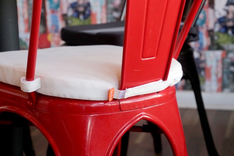 May include: A close-up of a red metal chair with a white cushion. The cushion is rectangular and secured with white straps. The chair is part of a set, with a black chair visible in the background.