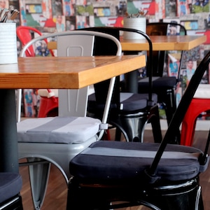 May include: A cafe interior featuring wooden tables and metal chairs in various colors. The chairs have padded seats with gray stripes. A metal container on the table holds silverware. The background has a colorful collage of images.