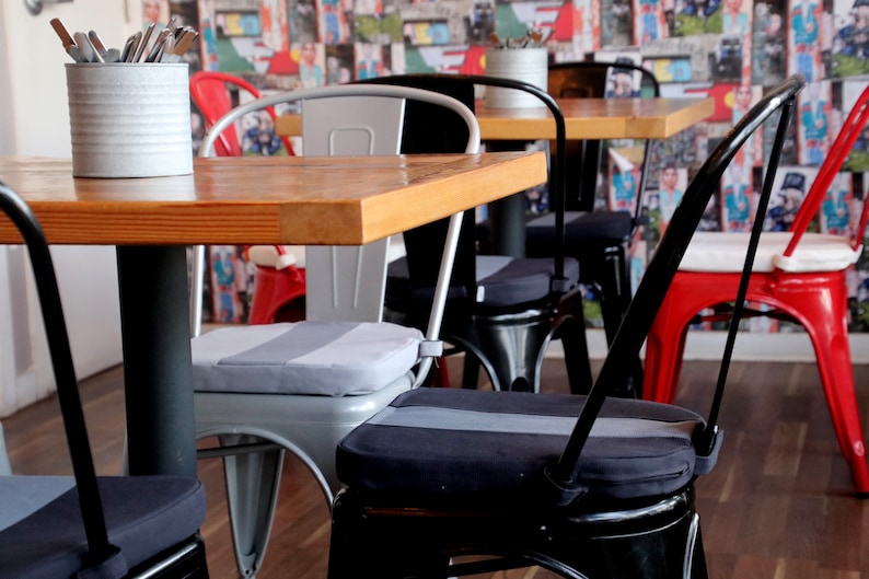 May include: A cafe interior featuring wooden tables and metal chairs in various colors. The chairs have padded seats with gray stripes. A metal container on the table holds silverware. The background has a colorful collage of images.