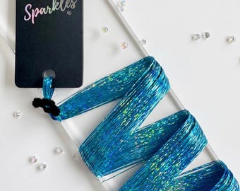 May include: A bundle of bright blue holographic hair tinsel strands attached to a black rectangular display card with the text Hair Sparkles printed in white and pink. The long, thin, metallic-finish strands are folded in a zigzag pattern.