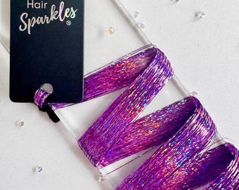 May include: A black rectangular display card with the text Hair Sparkles in white and iridescent lettering. Attached to the card is a bundle of vibrant purple holographic hair tinsel strands wrapped in a zigzag pattern around a clear acrylic base.