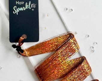 May include: A long, thin strand of copper-colored hair tinsel with a holographic, reflective finish. The tinsel is folded in a zigzag pattern and attached to a black rectangular display card labeled with the text Hair Sparkles in white.