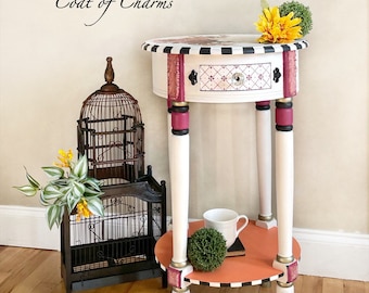May include: A white round end table with black and white trim, pink accents, and a single drawer. The table has a peach-colored top and gold accents on the legs. The table is decorated with a birdcage, a potted plant, and a cup and saucer.