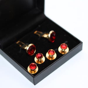 May include: A set of gold-coloured cufflinks and shirt studs with bright red gemstone details, displayed in a black box. The cufflinks and studs have a classic design, suitable for formal occasions.