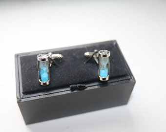 May include: A pair of silver cufflinks with a unique hourglass design. Each cufflink features a clear glass tube filled with blue sand, encased in a silver frame. The cufflinks are presented on a black velvet surface inside a black box.