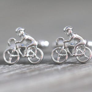May include: A pair of silver-toned metal cufflinks, each featuring a three-dimensional design of a cyclist riding a bicycle. The figures are depicted in a forward-leaning position, wearing helmets, and mounted on standard cufflink backings.