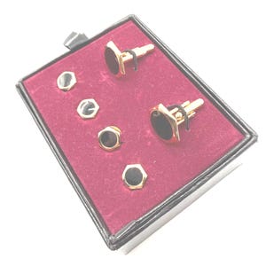 May include: A set of two gold-toned cufflinks and four matching shirt studs, each featuring a hexagonal shape with a smooth black inlay. The items are arranged on a deep red velvet-lined display box with a black exterior frame and a pull tab.