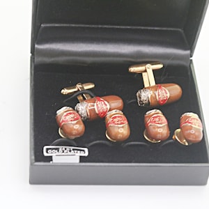 May include: A set of six 24ct gold plated cufflinks shaped like brown cigars with red and white labels, presented in a black box. The cufflinks feature a realistic cigar design with metallic gold hardware and a small rectangular tag reading 24KT GOLD PLATED.