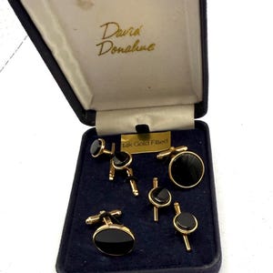 May include: A set of gold-filled cufflinks and shirt studs with circular black inlays, presented in a navy blue hinged box labeled David Donahue. The set includes two large cufflinks and four smaller shirt studs, all featuring a polished gold finish.