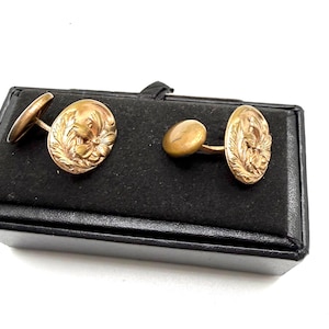 May include: A pair of oval-shaped gold-toned cufflinks featuring an embossed floral design with intricate leaf detailing. Each cufflink has a smooth, rounded toggle backing and a polished metallic finish, presented as a matching set.