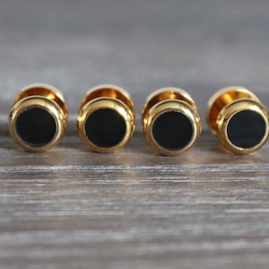 Pode incluir: Quatro botões de tachas circulares com uma borda de metal dourado polido que envolve uma incrustação central preta, lisa e plana. Cada botão tem uma base arredondada de cor dourada metálica e um design uniforme com um acabamento brilhante.