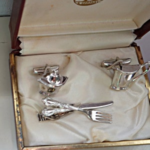 Peut inclure: Un ensemble vintage de boutons de manchette et d'attache de cravate argentés, avec des motifs miniatures de théière, de crémier et de couverts. L'ensemble est présenté dans un coffret marron et doré avec une doublure en satin crème, et la marque "DON" est visible.