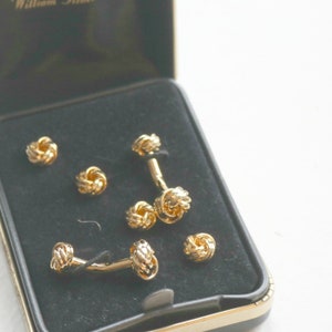 May include: A set of gold-toned knot cufflinks and shirt studs presented in a black box with a gold trim. The cufflinks and studs have a textured, woven design. The box is open, revealing the contents against a black velvet interior.