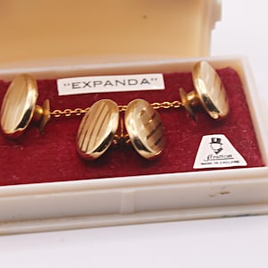 Könnte beinhalten: Goldfarbene Manschettenknöpfe mit ovalem Design, präsentiert in einem cremefarbenen Etui mit rotem Samtfutter. Die Manschettenknöpfe sind durch eine feine Kette verbunden und tragen den Text "EXPANDA". Ein kleines Etikett trägt die Aufschrift "Stratton Made in England".