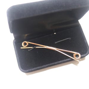 May include: A gold-toned metal collar bar with a straight pin design and circular ends, resting inside a rectangular black velvet-lined jewelry box. The accessory features a central sliding mechanism that connects the two thin metal bars.