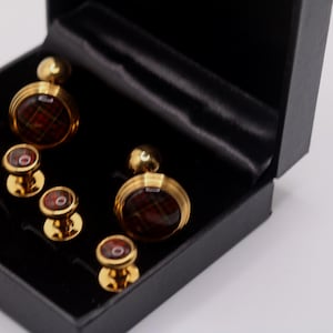 May include: A set of gold-toned cufflinks and shirt studs with a red and brown plaid design, presented in a black box with a satin lining. The set includes two pairs of cufflinks and three shirt studs.