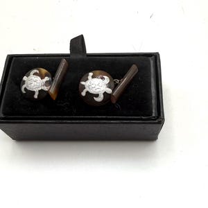 May include: A pair of round cufflinks featuring a silver-toned turtle motif set against a dark brown, mottled background. The cufflinks are presented in a black rectangular box with a black velvet interior lining and a small black fabric pull tab.