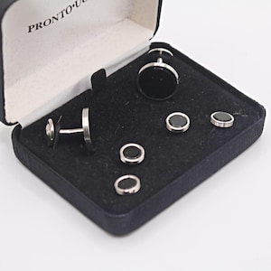 May include: A set of silver-toned metal cufflinks and shirt studs with circular black inlays, presented in a black hinged storage box with the text PRONTO UOMO printed on the interior lid. The set includes two cufflinks and four matching shirt studs.