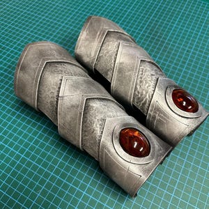 Pode incluir: Par de bra&ccedil;adeiras prateadas com desenhos geom&eacute;tricos e superf&iacute;cies texturizadas. Cada bra&ccedil;adeira apresenta uma grande gema oval, castanho-avermelhada. As bra&ccedil;adeiras s&atilde;o projetadas para proteger os antebra&ccedil;os.