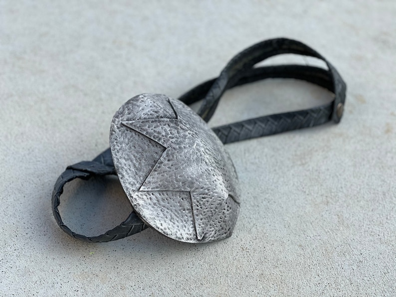 Pode incluir: Uma gravata bolo prateada com um pendente oval texturizado com um design de estrela. O pendente est&aacute; ligado a um cord&atilde;o de couro preto tran&ccedil;ado. Este acess&oacute;rio de estilo ocidental &eacute; uma joia cl&aacute;ssica.