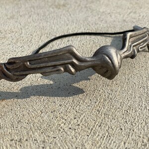 Pode incluir: Um acess&oacute;rio de cabelo de metal prateado com um design abstrato. A pe&ccedil;a apresenta uma forma estilizada e fluida com uma figura central e uma faixa el&aacute;stica preta presa. O acess&oacute;rio foi projetado para ser usado no cabelo.