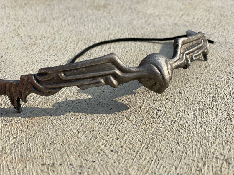 Pode incluir: Um acess&oacute;rio de cabelo de metal prateado com um design abstrato. A pe&ccedil;a apresenta uma forma estilizada e fluida com uma figura central e uma faixa el&aacute;stica preta presa. O acess&oacute;rio foi projetado para ser usado no cabelo.