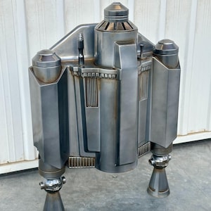 May include: A silver-colored metal model of a spaceship with a detailed design. The model has a rounded top and two legs. The model is weathered and has a distressed look.