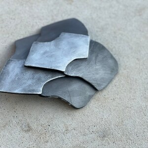 Pode incluir: Um conjunto de pe&ccedil;as met&aacute;licas sobrepostas de formas irregulares. As tonalidades de cinza variam, com &aacute;reas mais claras e outras mais escuras, criando um efeito de textura. As bordas s&atilde;o ligeiramente curvas.