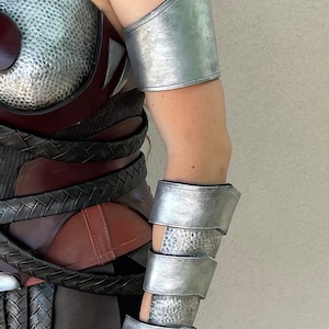 Pode incluir: Armadura de bra&ccedil;o prateada com textura martelada e uma gema vermelha. A armadura faz parte de uma fantasia, com um peitoral e uma saia a condizer. A fantasia tem um tema de guerreiro ou super-her&oacute;i.