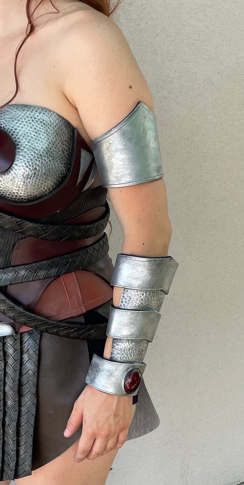 Pode incluir: Armadura de bra&ccedil;o prateada com textura martelada e uma gema vermelha. A armadura faz parte de uma fantasia, com um peitoral e uma saia a condizer. A fantasia tem um tema de guerreiro ou super-her&oacute;i.