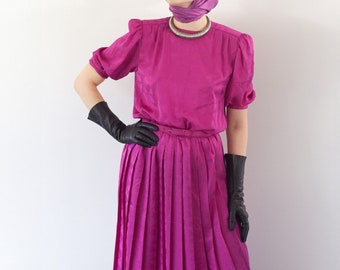 May include: A magenta dress with a pleated skirt and short sleeves. The dress has a fitted bodice and a wide waistband. The model is wearing black gloves.