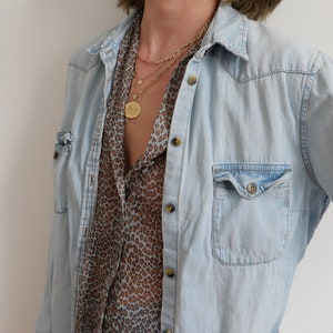 May include: A light blue denim shirt with a brown and white leopard print shirt underneath. The person is wearing a layered gold necklace with multiple pendants.