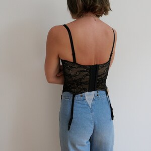 May include: Black lace corset with adjustable straps and a hook and eye closure in the back. The corset is worn over a pair of light blue denim jeans.