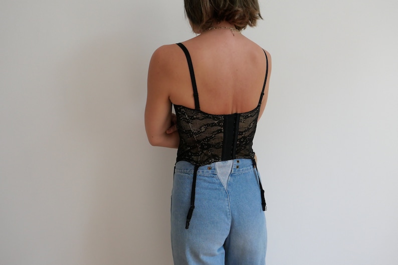 May include: Black lace corset with adjustable straps and a hook and eye closure in the back. The corset is worn over a pair of light blue denim jeans.