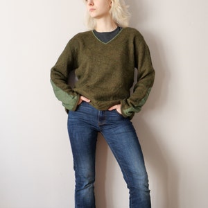 May include: A green sweater with a v-neck and green elbow patches. The sweater is made of a soft, fuzzy material. The person wearing the sweater is also wearing blue jeans.