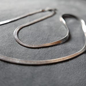 Può includere: Una collana a catena di serpente argentata, con due fili di catena, su una superficie di tessuto grigio.