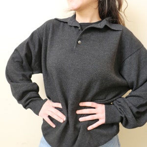 May include: A dark gray long-sleeved sweater with a collared neckline and a single button closure. The sweater has a relaxed fit and ribbed cuffs. The model is wearing blue jeans.