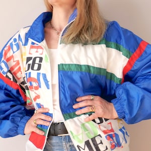 May include: A retro-style jacket in a striking colour palette. The jacket is predominantly blue, with white, green, and red detailing. The front panels display a colourful print with letters and numbers. It has a zip-up front and a high collar.