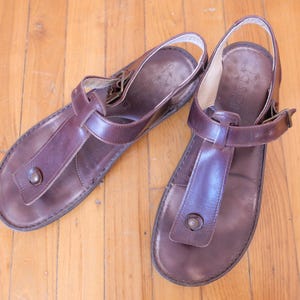 May include: A pair of brown leather sandals with a thong-style toe strap and ankle strap. The sandals have a buckle closure and a small metal button detail on the toe strap. The sandals are on a wooden floor.