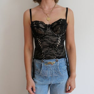 May include: A black lace corset with boning and adjustable straps. The corset has a sweetheart neckline and is worn over a pair of light blue jeans.