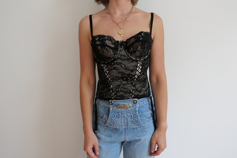 May include: A black lace corset with boning and adjustable straps. The corset has a sweetheart neckline and is worn over a pair of light blue jeans.