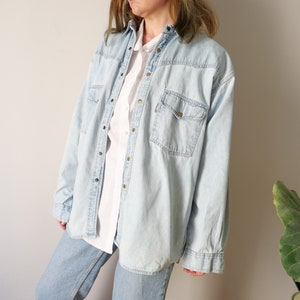 Pode incluir: Uma camisa de ganga azul claro com botões e mangas compridas. A camisa tem um bolso no peito e um caimento descontraído.