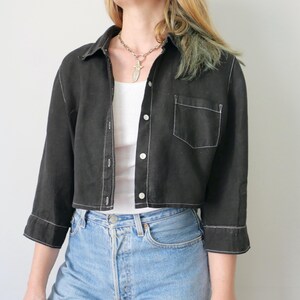 May include: Black cropped button-down shirt with white stitching detail. The shirt has a collared neckline, a single chest pocket, and three-quarter sleeves. The shirt is paired with light blue denim jeans.