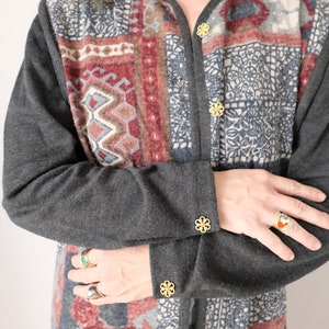 May include: A grey cardigan with a colourful patterned design. The cardigan has gold buttons and is worn with the sleeves rolled up.