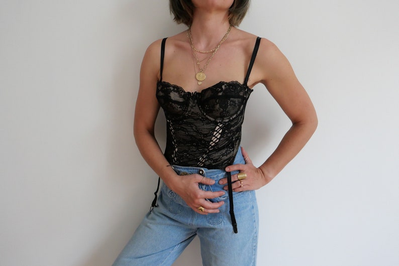 May include: A black lace corset with a sweetheart neckline and adjustable straps. The corset is worn over a pair of light blue jeans.
