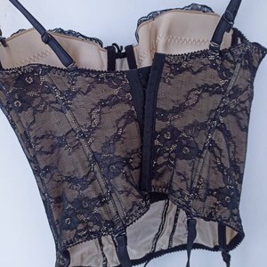 May include: Black lace corset with beige lining and adjustable straps. The corset has a hook and eye closure in the back and garter straps at the bottom.