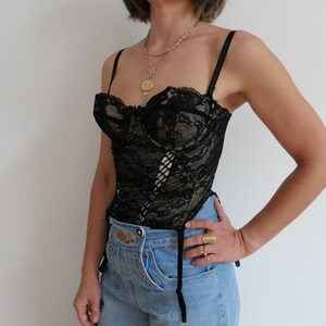 May include: A black lace corset with a lace-up front and adjustable straps. The corset is worn over a pair of light blue jeans.