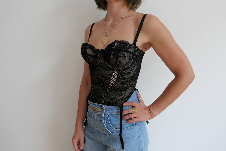 May include: A black lace corset with a lace-up front and adjustable straps. The corset is worn over a pair of light blue jeans.